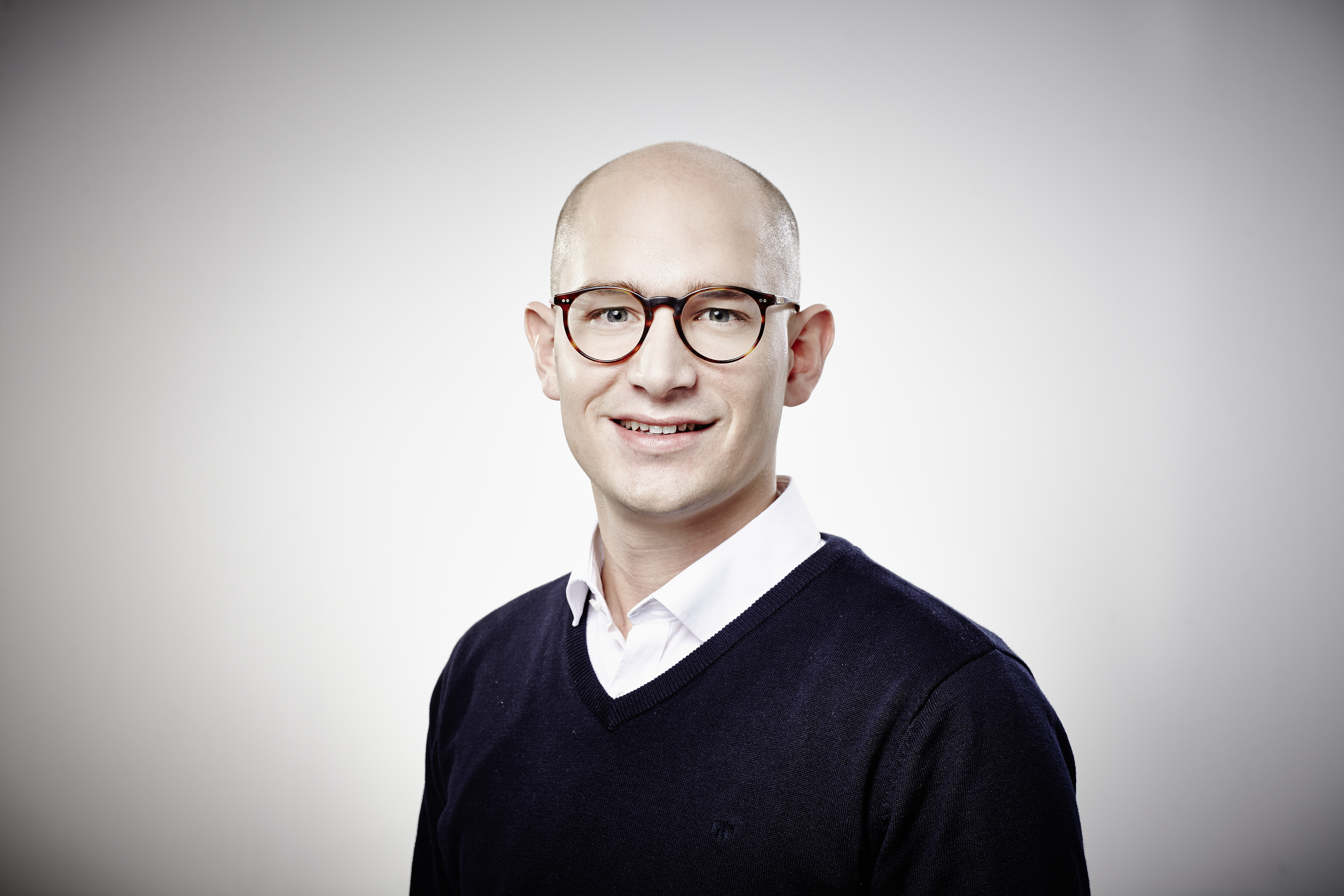 Oliver Schopp mit einem leichten Lächeln vor einem grauen Hintergrund. Er trägt sehr kurze Haare, eine große braune Brille und ein weißes Hemd unter einem dunklen V-Ausschnitt-Pullover.